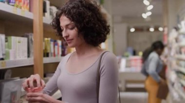 Makyaj dükkanında dikilen kıvırcık saçlı, beyaz bir kızın orta boy yakın çekimi nemlendirici krem kavanozunun kapağını açıyor, kokluyor, aromanın tadını çıkarıyor, kafa sallıyor, gülümsüyor ve rafa koyuyor.