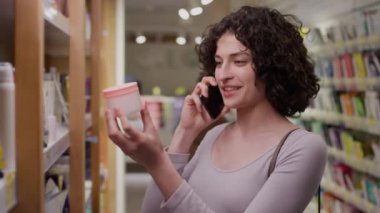 Heyecanlı beyaz kadın müşterinin yeni güzellik mağazasında durup raflardan kavanoz ve şişe dolusu cilt ürünleri alıp etiketlere bakarken, akıllı telefondan kız arkadaşıyla sohbet ederken orta boy görüntüleri.