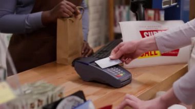 Genç Afro-Amerikan satış asistanının kasada müşteriye servis yaparken orta ölçekli çekimi. Terminalde kart ile ödeme yapan kimliği belirsiz bir kadın. Gülümseyen, teşekkür eden ve alışveriş torbasını teslim eden kadın.