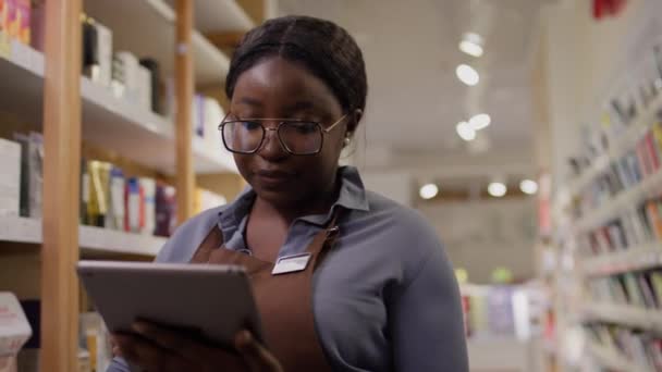 身着眼镜和围裙的非洲裔美国女化妆品店年轻员工站在商店地板上 清点货架上的商品库存 并定购新用品的中等镜头 — 图库视频影像