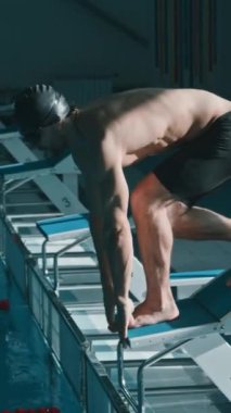 Şortlu, şapkalı, gözlüklü genç erkek yüzücünün tam dikey görüntüsü dalış bloğunda duruyor ve mükemmel bir atlayış yapıyor, sonra mavi suyun altında kayboluyor, havuz eğitimi sırasında.