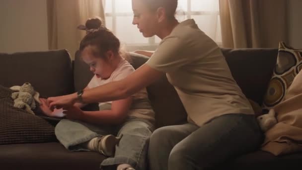デジタルタブレットを使用して自宅で漫画を見ているダウン症の娘と座っている母親の側面映像 — ストック動画