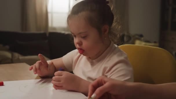 无法辨认的保姆或母亲教患有抑郁症的白人小女孩在家里坐在桌旁用蜡笔画画的侧影 — 图库视频影像