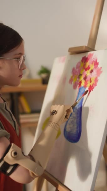 メガネと赤いジャンプスーツで落ち着いた白人少女の垂直側の映像は 自宅で人工的な腕でゆっくりと花を描きます — ストック動画