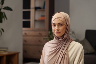 Oturma odasında oturmuş kameraya bakan, tesettürlü zarif Müslüman kadının orta boy yakın çekimi.