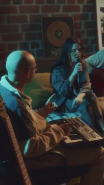Akşamları retro eski okul stüdyosunda üç rock şarkısını kaydeden müzik grubunun dikey yan görüntüleri. Vokalist ve ritimciye eşlik eden gitarist Midi kontrolörüyle çalıyor