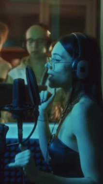 Kafkasyalı kadın vokalistin dikey yavaşlaması kayıt kabininde mikrofona imza atarken müzik yapımcısı stüdyoda kaydı kontrol ediyor.