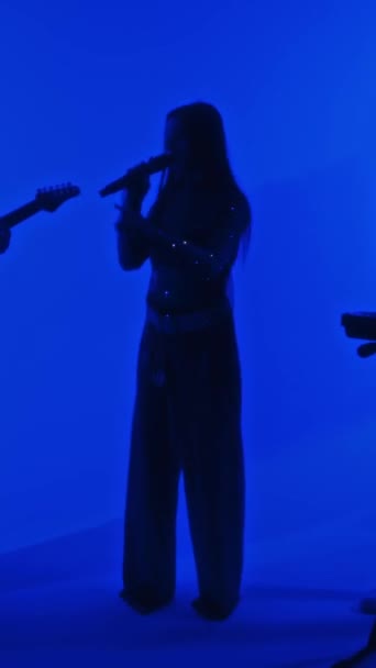 ブルーカラーのネオンフィルターでプロのスタジオでギグする女性歌手とミュージシャンのミュージカルトリオの垂直フルビデオ — ストック動画