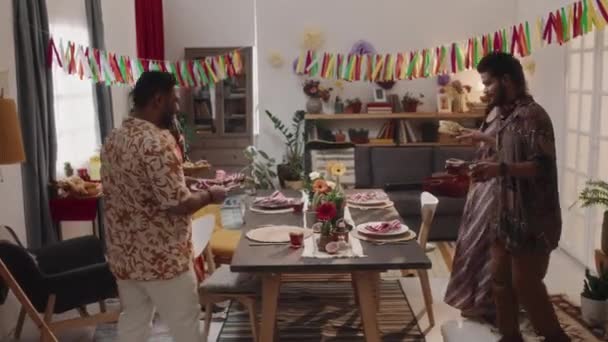 明るいカラフルなシャツとドレスの4人の若いヒスパニック系男性と女性のフルズーム映像は 伝統的なメキシコの食べ物とカクテルを持って豪華な休日の饗宴のためにテーブルに持って来ます — ストック動画