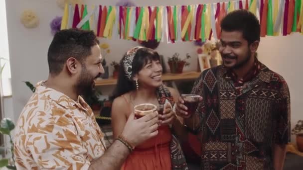 カラフルなシャツと髪のスカーフを持つ女性の2人の若いメキシコ人またはヒスパニックの男性のメディア映像 家のパーティーでガーランドの下に立っている明るいドレス チャット カクテルを楽しむ — ストック動画