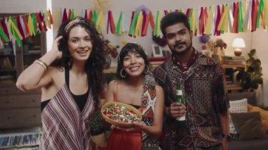 Üç Meksikalı 'nın orta boy portresi, renkli elbiseli iki kız, elinde bir kase salata ve elinde bira olan sakallı bir adam, kurdele altında eğlenceli fotoğraflar için poz veriyor.