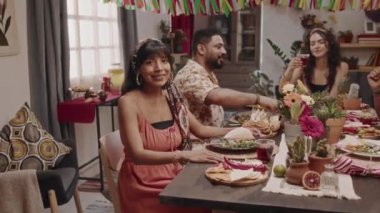 Cinco de Mayo partisi sırasında yemek masasında oturan, kameraya gülümseyen, arka planda kokteyllerle kadeh kaldıran genç ve güzel Meksikalı kadının orta boy portre görüntüsü.