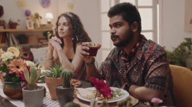 Orta boy sakallı ve bıyıklı genç Meksikalı adam, desenli gömlekli, elinde alkollü içki, festival masasında oturuyor, arkadaşlarıyla konuşuyor, karısını dinliyor ve izliyor.