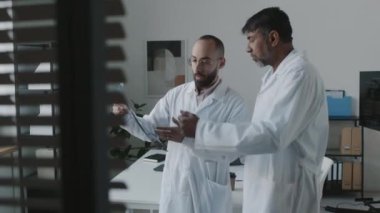 Orta Doğu 'lu beyaz önlüklü bir erkek klinisyenin hastanedeki Asyalı başhekime zor bir hasta vakası hakkında danışmanlık yapması, röntgene bakması, anormalliklere işaret etmesi.