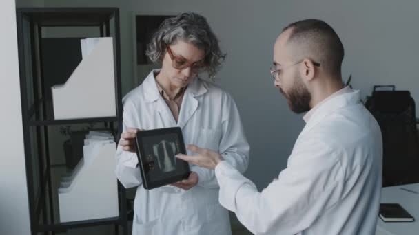 身穿白衣的成熟白种人女医生和中东男性同事一起站在办公室里 拿着带有X光图像的平板电脑 讨论罕见的医疗案例的中等镜头 — 图库视频影像