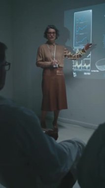 Gözlüklü, bluzlu, kimlik kartlı, orta yaşlı beyaz bir kadının tam dikey çekimi. Toplantıda projektör ekranda duruyor. Doktorlara biyokimya üzerine bilimsel araştırmaları anlatıyor.