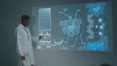 Yetişkin Hintli bilim adamı ya da doktorun sahnede projektör ekranının yanında durup, mikrobiyoloji üzerine yeni araştırmalar hakkında ders verirken, hücre çizimleriyle slayta işaret ederken ve seyircilere konuşurken.