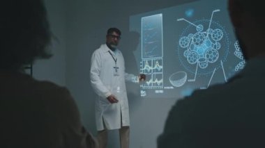 Yetişkin Asyalı erkek araştırmacının sahnede projektör ekranının yanında durup, immünolojide yeni bilimsel keşifler hakkında konuşurken, hücre çizimlerini gösterirken, halkla konuşurken,