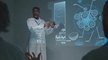 Orta boy Hintli erkek doktor sahnede projektör ekranın yanında duruyor, bölüm toplantısında yeni ilaç ya da terapinin keşfi hakkında konuşuyor, selamlıyor ve meslektaşlarını alkışlıyor.