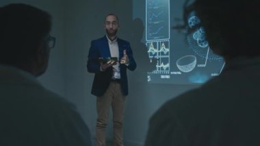 Orta Doğulu erkek araştırmacı bilim adamının tıp konferansında beyaz önlüklü doktorlara yeni ilacın yenilikçi keşfi, ekranda slayt gösterisi, seyirci alkışlaması...