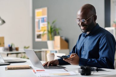 Modern ofiste akıllı telefon kullanan ve dizüstü bilgisayarda yazan neşeli Afro-Amerikan yöneticinin orta boy görüntüsü.