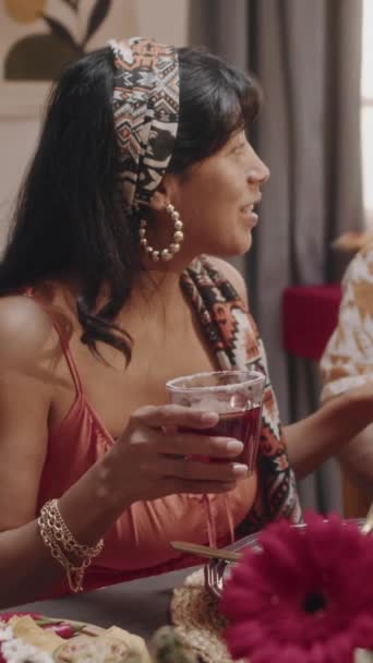 髪に明るいスカーフを持つ美しいメキシコの若い女性のミディアム垂直映像 パーティーテーブルに座って夏のドレスで マルガリータカクテルを開催 友達に面白い話を伝える — ストック動画