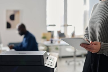 Modern ofiste dijital tablet kullanarak çalışan tanınmayan genç bir kadının seçici odak noktası. Kağıt belgeler basarken, boşluk kopyalarken.