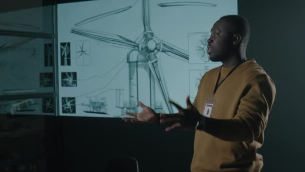 男性のアフリカ系アメリカ人グリーンエネルギーエンジニアのサイド映像は 会議中にオーディエンスにレイアウトで風力タービンローターブレードの生産を進めることについての彼のプロジェクトを提示します — ストック動画