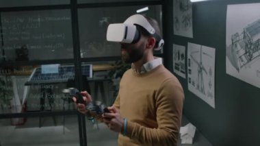 Gece yenilenebilir enerji ofisindeki güneş panelleri ve rüzgar türbinlerini görselleştirirken, erkek sakallı mühendisin VR kulaklık ve kontrolörleri kullanışının yan görüntülerini de ekleyin.