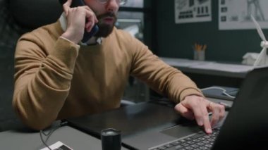 Sakallı, çift taraflı yeşil enerji uzmanının dizüstü bilgisayarla çalışan ve gece ofis masasında otururken akıllı telefonla konuşan görüntüsü.