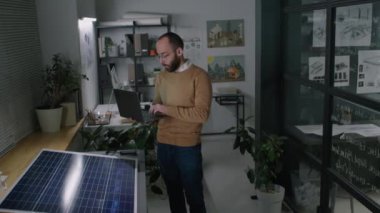 Erkek güneş enerjisi mühendisinin güneş paneli parçasının yanında duran ve iş günü tavan arasında dizüstü bilgisayara bilgi ve özellikleri hızla yazan tam bir yan görüntüsü.