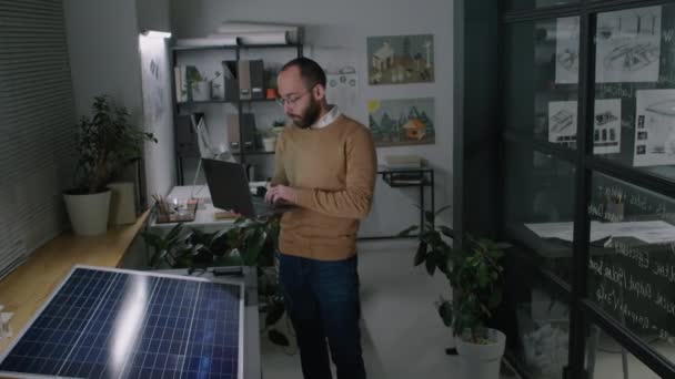 在阁楼办公室工作期间 男性太阳能工程师站在太阳能电池板碎片附近 在笔记本电脑上快速输入信息和特征的完整媒体画面 — 图库视频影像