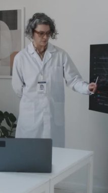 Orta yaşlı, beyaz önlüklü, hastane personeli toplantısında konuşan beyaz önlüklü beyaz kadın tıbbi direktörün orta dikey görüntüsü, kazanç ve gelir bildirimi, meslektaşlarının sorularını cevaplamak.