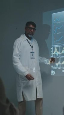 Yetişkin Asyalı erkek bilim adamının tam dikey görüntüleri projektör ekranın yanında duruyor, yenilikçi tedavi sunuyor, konuşuyor ve grafikler ve çizelgeler ile slayta işaret ediyor.