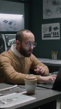 Masada dizüstü bilgisayarda yazan gözlüklü erkek melez mühendisin dikey yan görüntüsü. Geceleri duvarların planlarıyla yenilenebilir enerji kaynakları geliştirmeye çalışırken.