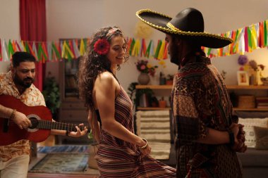 Genç neşeli esmer kadın partide müzisyen akustik gitar çalarken İspanyol adamın önünde şapkalı dans ediyor.