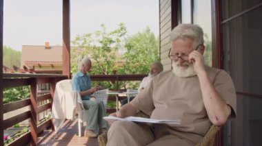 Emeklilik cemiyetinin terasında oturan, elinde gazeteyle oturan, kameraya gülümseyen, arka planda sohbet eden çiftlerin orta ölçekli portre görüntüleri.