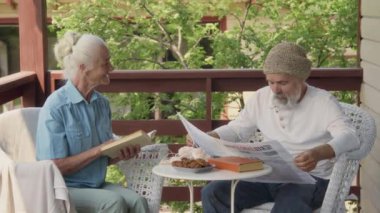 Orta sınıf kıdemli beyaz kadın ve erkek sabah terasta oturuyor, koca gazete okuyor, karısı kitap okuyor, konuşuyor ve haber makalesine işaret ediyor.