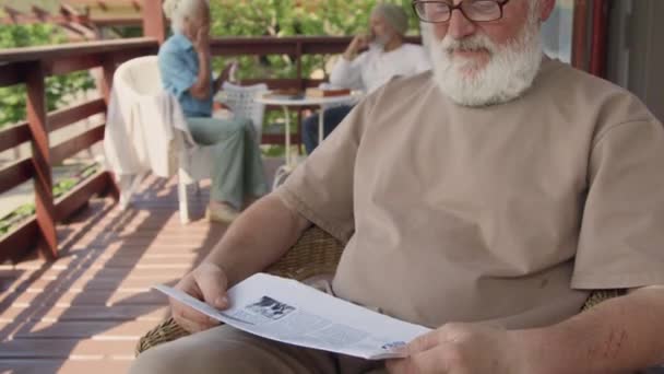70歳の白人男性が灰色の髪をした中型のタイル張りショット 引退した家のテラスのアームチェアに座り 新聞を読み メガネを外し カメラを見て笑顔で — ストック動画