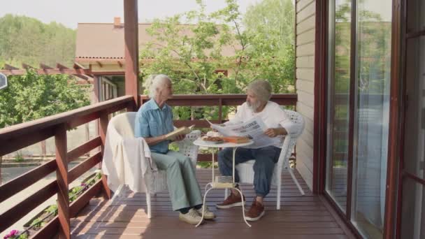 ウッリー帽子の高齢の白人男性のワイドショット 新聞を読み 晴れた夏の日の退職の家のテラスに座って本を保持している脆弱な女性とニュースを議論 — ストック動画
