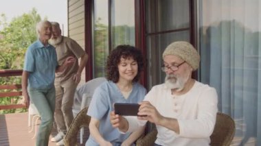 Üst düzey beyaz adam ve hemşirenin terasta yan yana oturduğu orta büyüklükte görüntüler birlikte poz vermeleri, gülümsemeleri ve akıllı telefondan selfie çekmeleri için çağrıda bulunuyorlar.