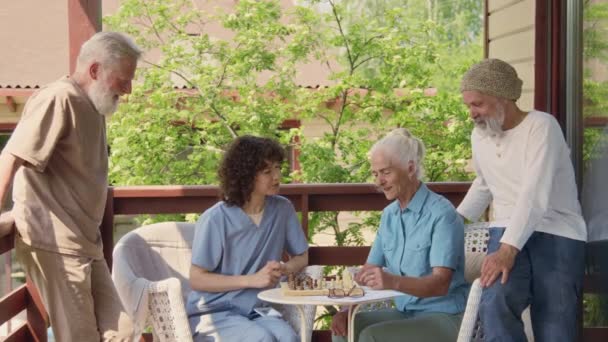青い制服の笑顔の若い女性看護師と退職後のコミュニティのテラスでチェスをする高齢の白人女性 次の動きを計画し 男性の友人にアドバイスを求める — ストック動画