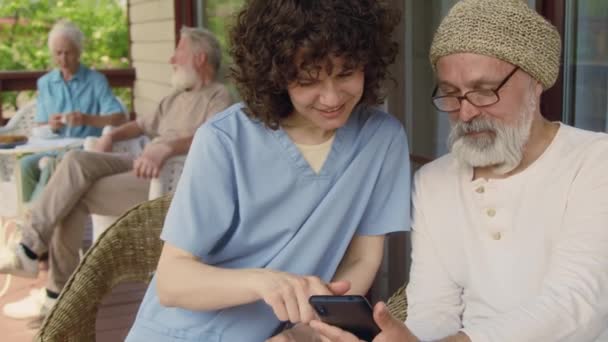 青い制服の陽気な女性看護師と高齢の白人男性が引退した家のテラスのアームチェアに座って スマートフォンでインターネットを閲覧することを学びます — ストック動画