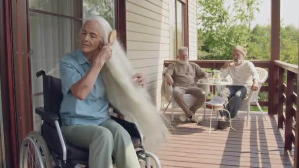 テラスの屋外の車椅子に座り 長い銀髪をブラッシングし テーブルでチャットする美しい80以上の白人女性退職コミュニティのメディアフルビデオ — ストック動画