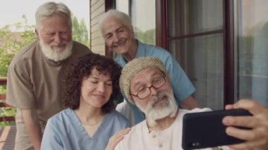 Yaşlı erkek ve kadın Kafkasyalı emekliler topluluğunun orta boy yakın çekimi ve terasta birlikte poz veren gülümseyen hemşire, akıllı telefondan eğlenceli selfie çekmesi ve sonucu kontrol etmesi.