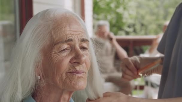 美しい80歳の白人女性のクローズアップ映像は 彼女の髪を穏やかにブラッシングしている制服で タトゥー付きの手で認識できない女性看護師にチャットしながら笑顔を浮かべます — ストック動画