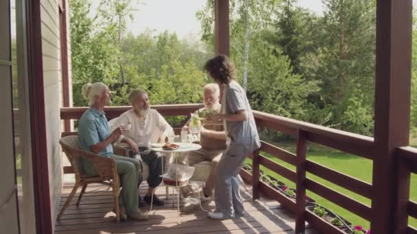 老年男女退休人士在阳台上围坐在桌旁 快乐的年轻护士带着下午的小吃和点心 与病人聊天 然后离开 — 图库视频影像