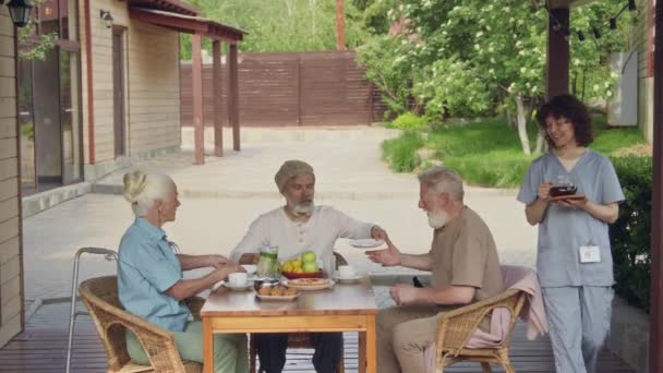 身着蓝色制服 满脸笑容的女护士 为退休后坐在户外的白种人端着一壶茶 围坐在桌子上 邀请吃饭 拍拍肩膀 — 图库视频影像
