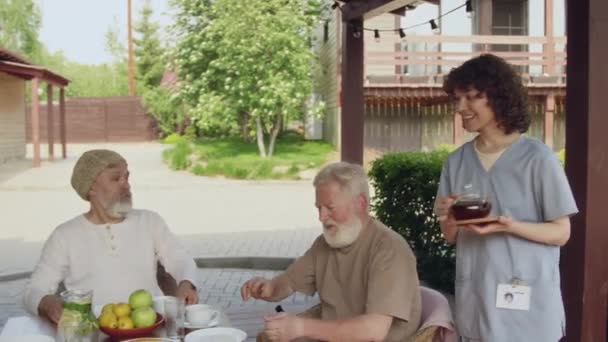 庭で屋外に座って おしゃべりして離れ 女性のコンパニオンに紅茶を注いでいる先輩の家の住民のための均一な陽気な看護婦のミディアム映像 — ストック動画