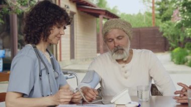 Orta ölçekli, üniformalı genç bayan hemşire ve dışarıda masada oturan yaşlı beyaz erkek hasta, kan basıncını manşet monitörüyle ölçüyor, tartışıyor ve sonucu yazıyor.